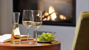 Zwei befüllte Weingläser stehen neben einer Karaffe und Trauben auf einem Tisch vor einem Kamin