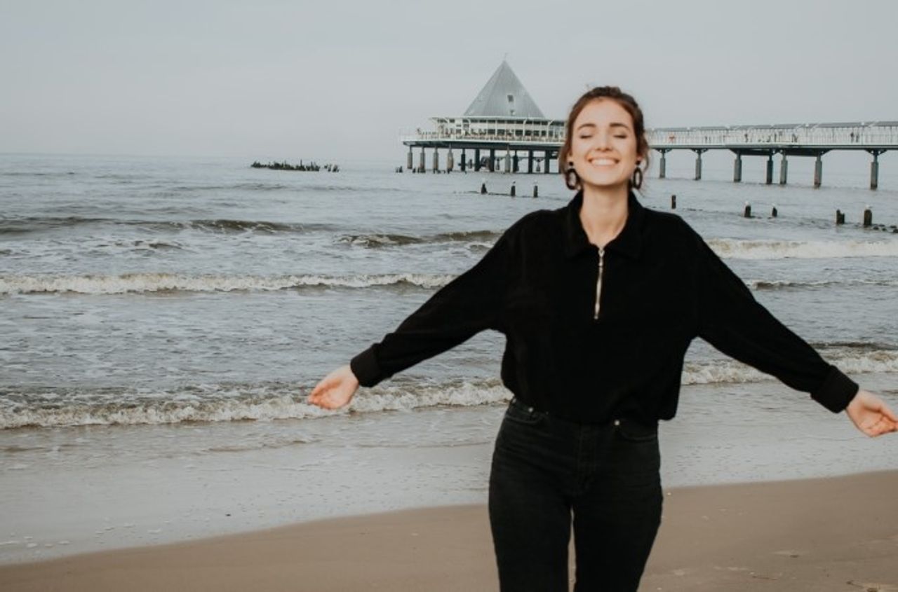 Foto von einer jungen Dame im Sommerurlaub in Heringsdorf, die ihre Arme ausbreitet und lächelt, im Hintergrund sieht man das Meer